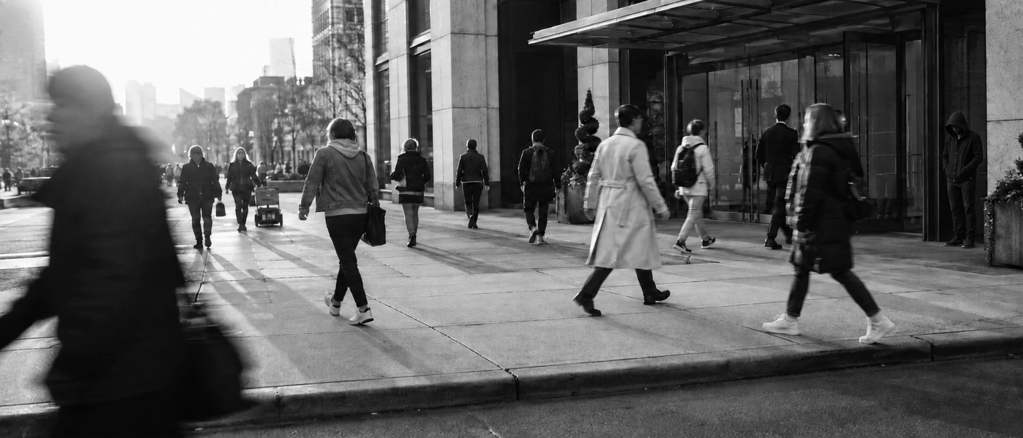 Pedestrians walking toward an office building entrance on a city sidewalk in early morning light